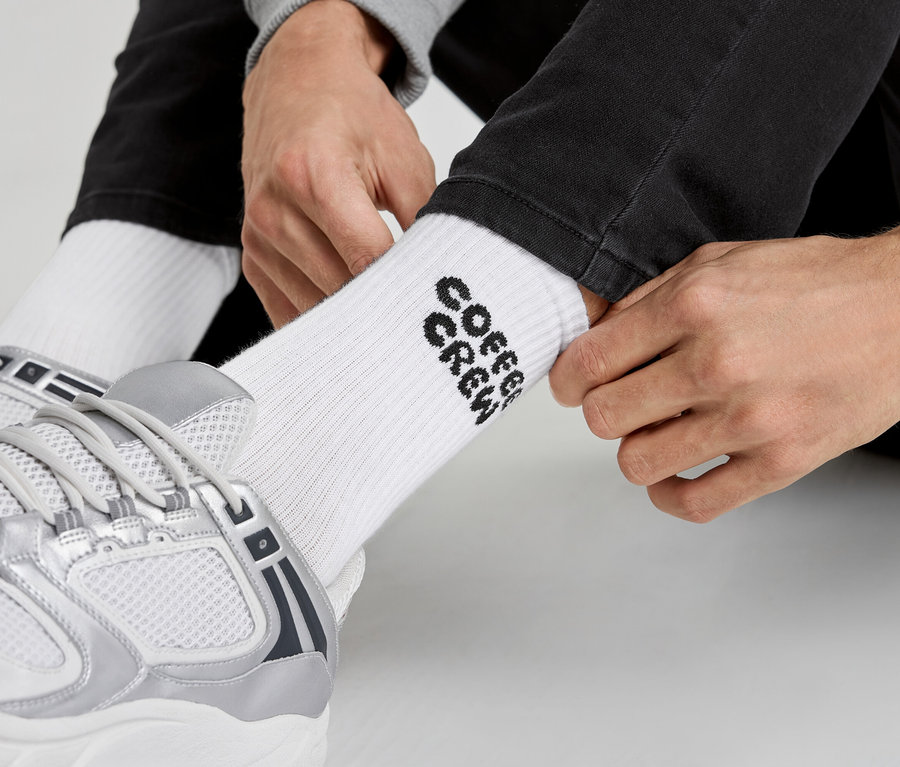 Un homme enfile des chaussettes blanches avec l'inscription « Coffee Crew » et des baskets argentées.