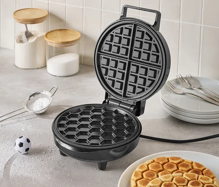 Gaufrier noir ouvert sur un comptoir de cuisine avec des gaufres finies dans une assiette, un petit ballon de football et des ingrédients dans des pots en verre en arrière-plan.
