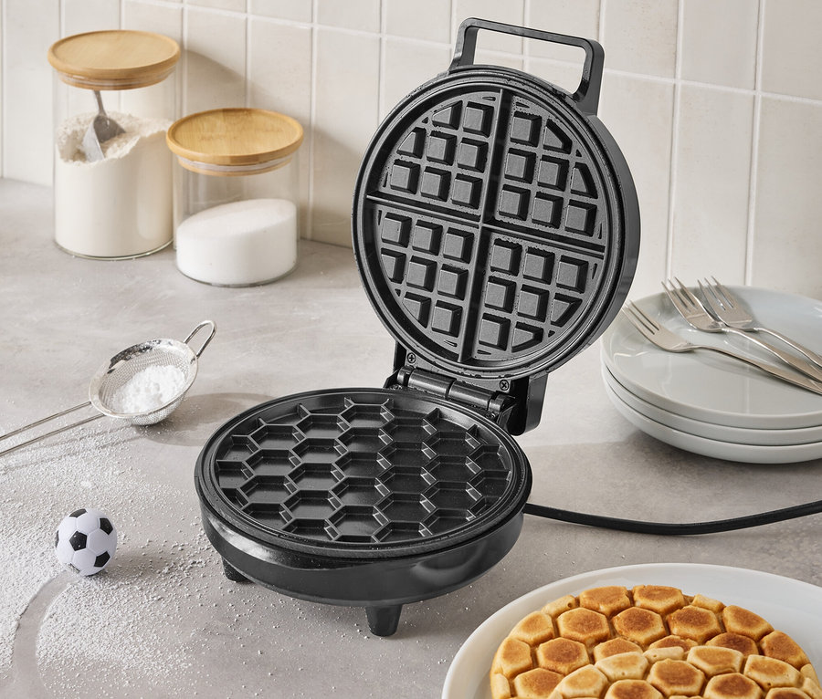 Gaufrier noir ouvert sur un comptoir de cuisine avec des gaufres finies dans une assiette, un petit ballon de football et des ingrédients dans des pots en verre en arrière-plan.