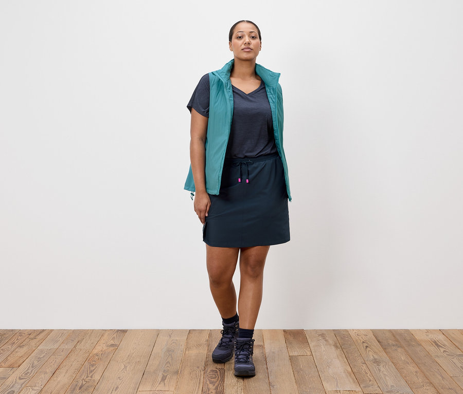 Une femme se tient devant un mur blanc, vêtue d'une jupe bleu marine, d'un t-shirt bleu marine, d'un gilet bleu pétrole et de chaussures de randonnée bleu marine.