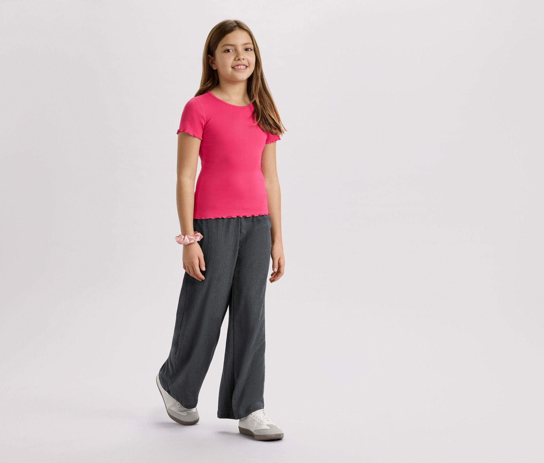 Une fille porte un t-shirt rose, un pantalon gris et des baskets sur un fond blanc.