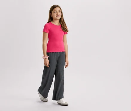 Une fille porte un t-shirt rose, un pantalon gris et des baskets sur un fond blanc.