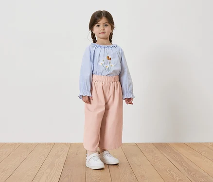 Fillette avec des tresses, portant un chemisier bleu clair avec des broderies florales, un pantalon rose et des baskets blanches.