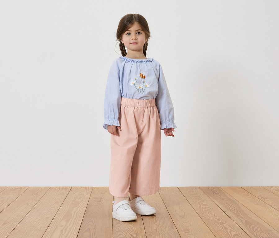 Fillette avec des tresses, portant un chemisier bleu clair avec des broderies florales, un pantalon rose et des baskets blanches.