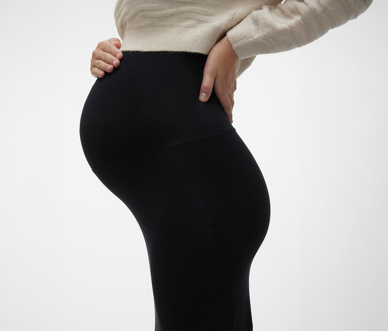 Femme enceinte portant une jupe maxi noire MAMALICIOUS «
MLEMMA TUBE
» et un pull beige, les mains sur le ventre.