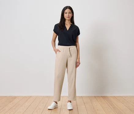 Une femme se tient devant un mur blanc. Elle porte un t-shirt noir, un pantalon beige et des baskets blanches.