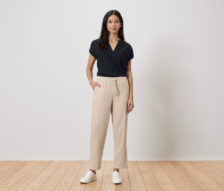 Une femme se tient devant un mur blanc. Elle porte un t-shirt noir, un pantalon beige et des baskets blanches.