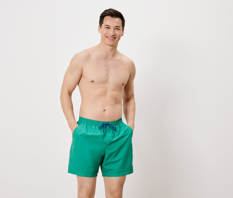 Homme torse nu en short de bain vert avec cordon bleu, les mains dans les poches, sur fond blanc.