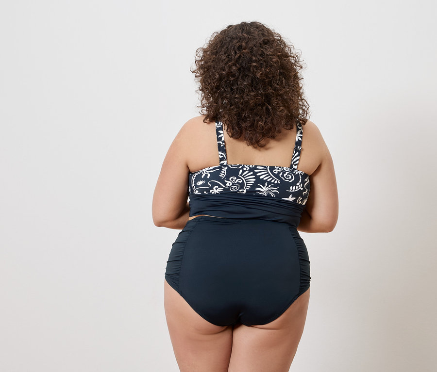 Vue arrière d'une femme aux cheveux bouclés portant un haut de tankini bleu marine à motifs blancs et un bas de bikini taille haute bleu marine.
