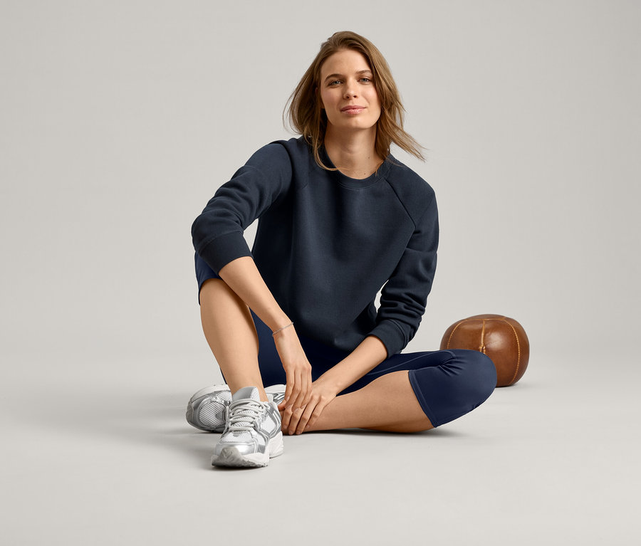 Une femme est assise en tailleur, portant un sweat-shirt bleu marine, des leggings bleu marine et des baskets argentées.
