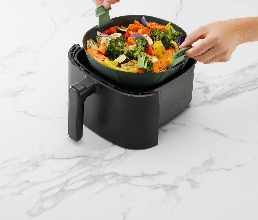 Une personne place un récipient vert rempli de légumes dans une friteuse à air noire sur une surface en marbre.