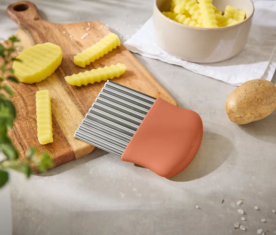 Une lame à frites ondulée repose sur une planche à découper avec des pommes de terre et des frites, à côté d'un bol de frites.