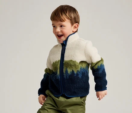 Un jeune garçon aux cheveux bruns, portant une veste polaire color-block blanche, verte et bleue et un pantalon vert, sourit et regarde vers la droite.