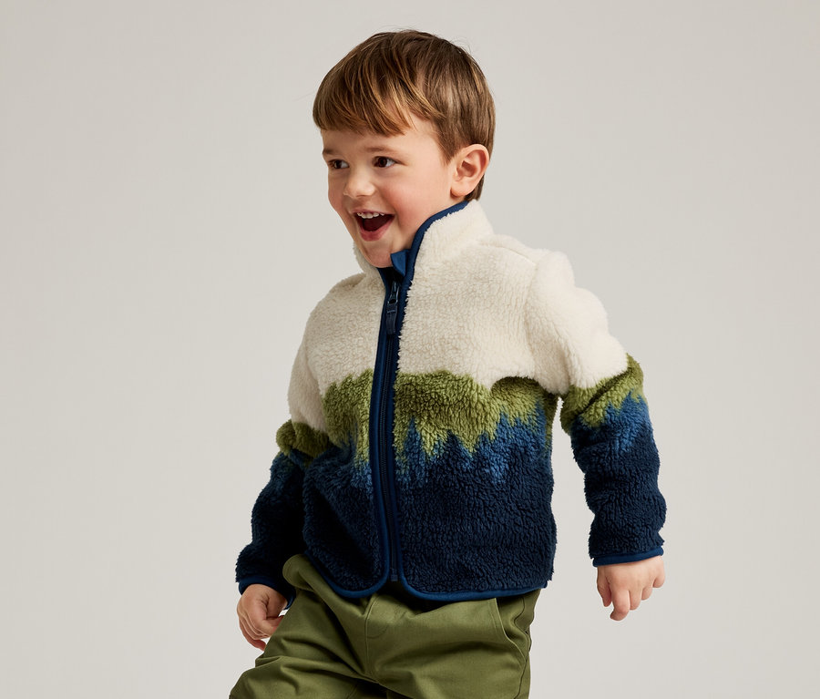 Un jeune garçon aux cheveux bruns, portant une veste polaire color-block blanche, verte et bleue et un pantalon vert, sourit et regarde vers la droite.