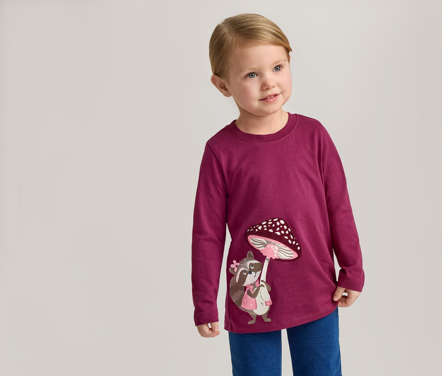 Un enfant blond porte un t-shirt à manches longues bordeaux avec un raton laveur et un champignon dessus, et un pantalon bleu.