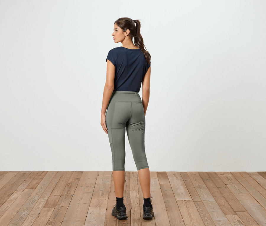Femme avec queue de cheval, vue de dos, portant un t-shirt bleu marine et un legging capri vert olive devant un mur blanc.