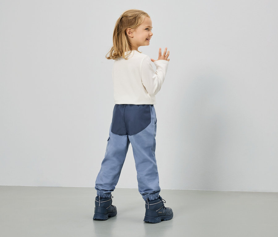 Une petite fille blonde vêtue d'un T-shirt blanc à manches longues, d'un pantalon bleu et de bottes bleu marine se tient devant un mur blanc.