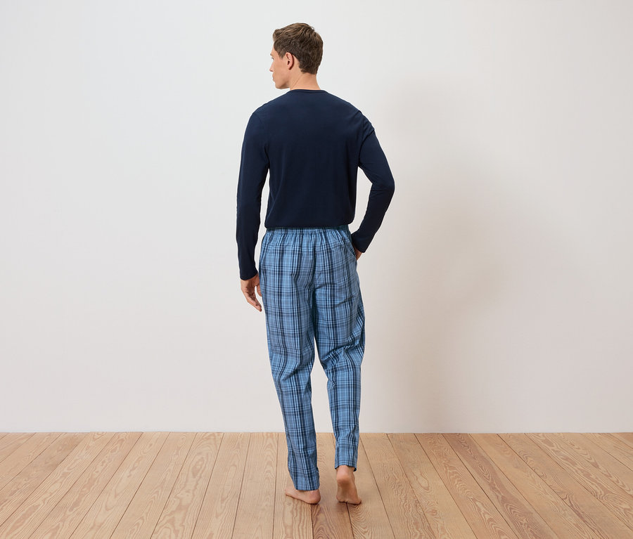 Homme vu de dos portant un pantalon de pyjama à carreaux bleus et un haut à manches longues devant un mur blanc sur un plancher en bois.