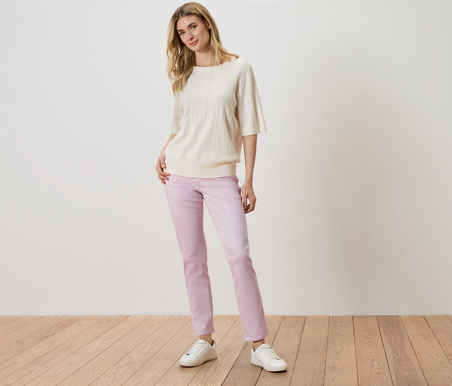 Une femme debout devant un mur blanc, portant un pull en tricot crème, un pantalon violet et des baskets blanches.