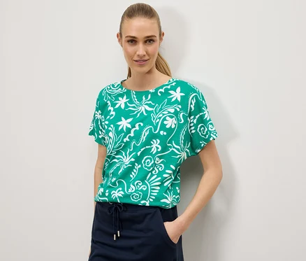 Une femme porte un t-shirt vert avec un motif floral blanc et une jupe bleu marine sur un fond blanc.