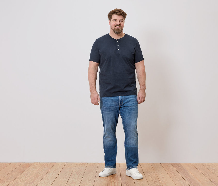 Un homme se tient devant un mur, il porte un t-shirt bleu, un jean et des chaussures blanches.