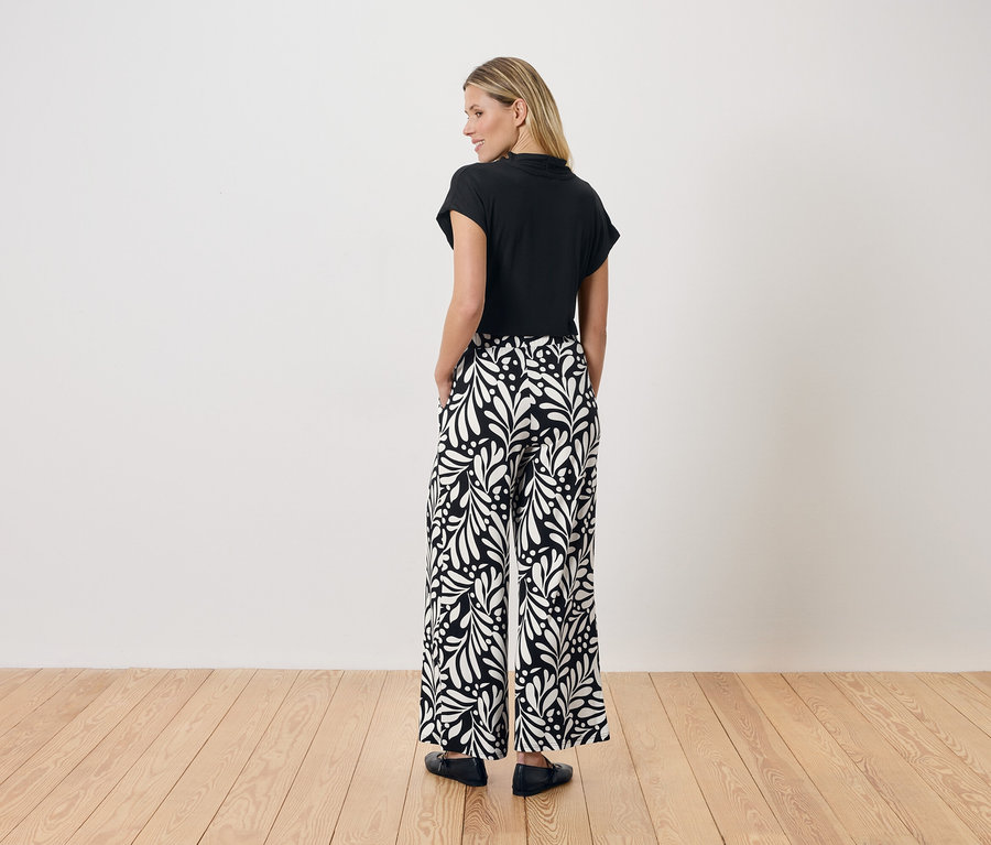 Une femme se tient dos à la caméra, portant un haut noir et un pantalon ample avec un motif de feuilles noir et blanc.