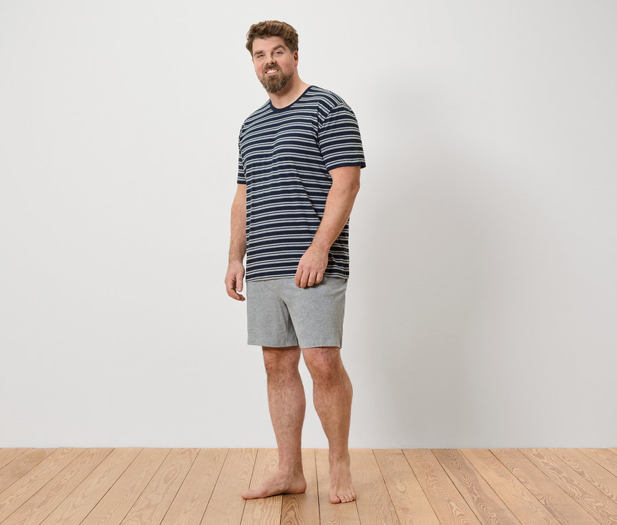 Un homme se tient pieds nus sur un plancher en bois, portant un t-shirt rayé et un short gris.