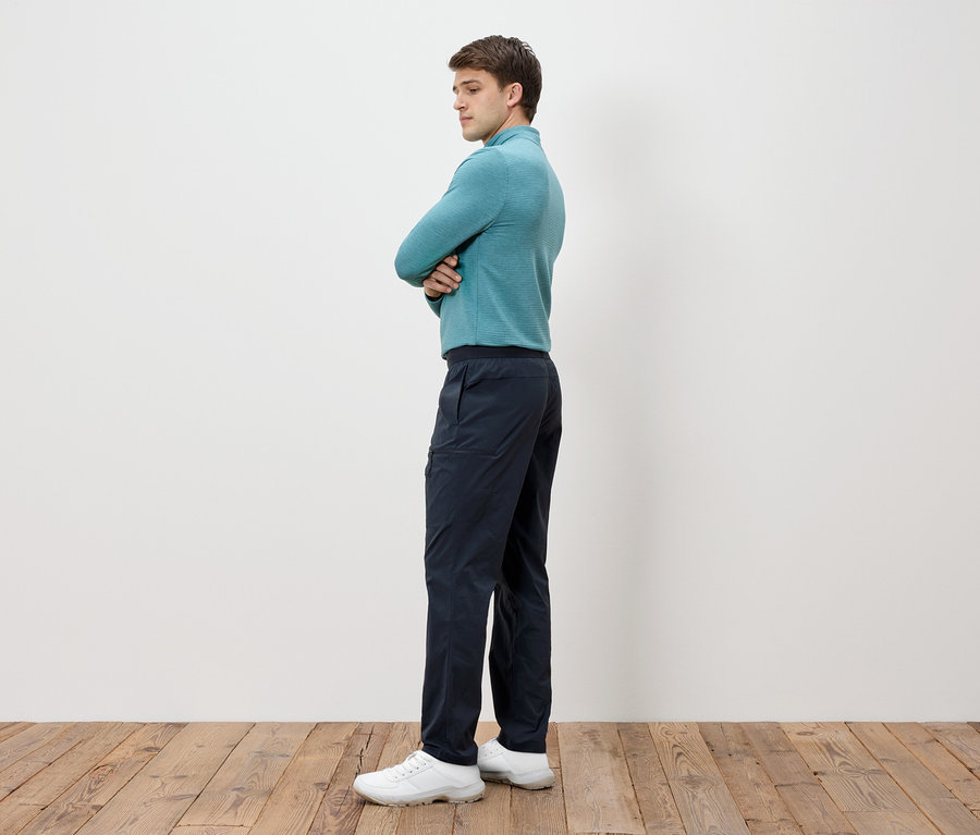 Homme les bras croisés, portant un t-shirt à manches longues bleu-vert, un pantalon foncé et des chaussures blanches.