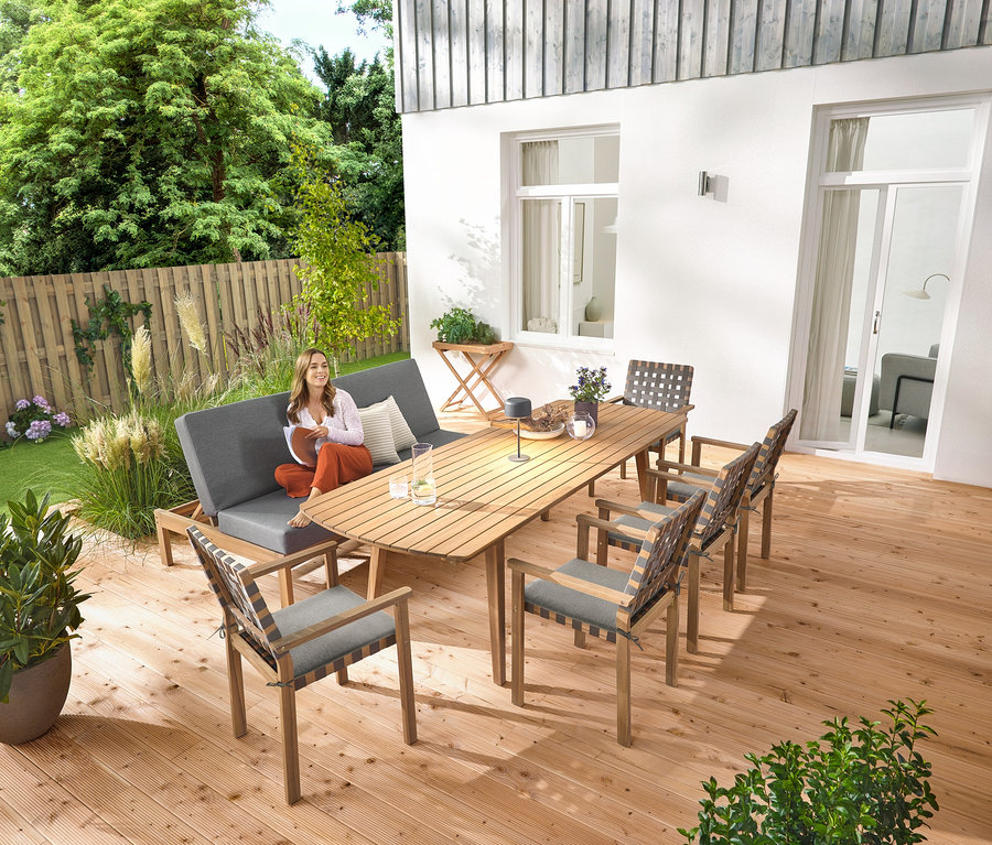 Une femme est assise sur un banc de jardin 2 en 1 ‒ double bain de soleil. Une lampe de table solaire grise est posée sur la table de jardin extensible avec des façades arrondies d'environ 180‒240 cm. Il y a aussi une table de jardin et des chaises de jardin empilables avec un tressage de ceinture.