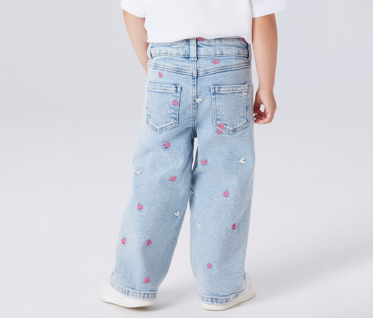 Enfant vu de dos portant un jean à jambes larges pour enfant NAME IT avec motif de cœurs et des chaussures blanches.