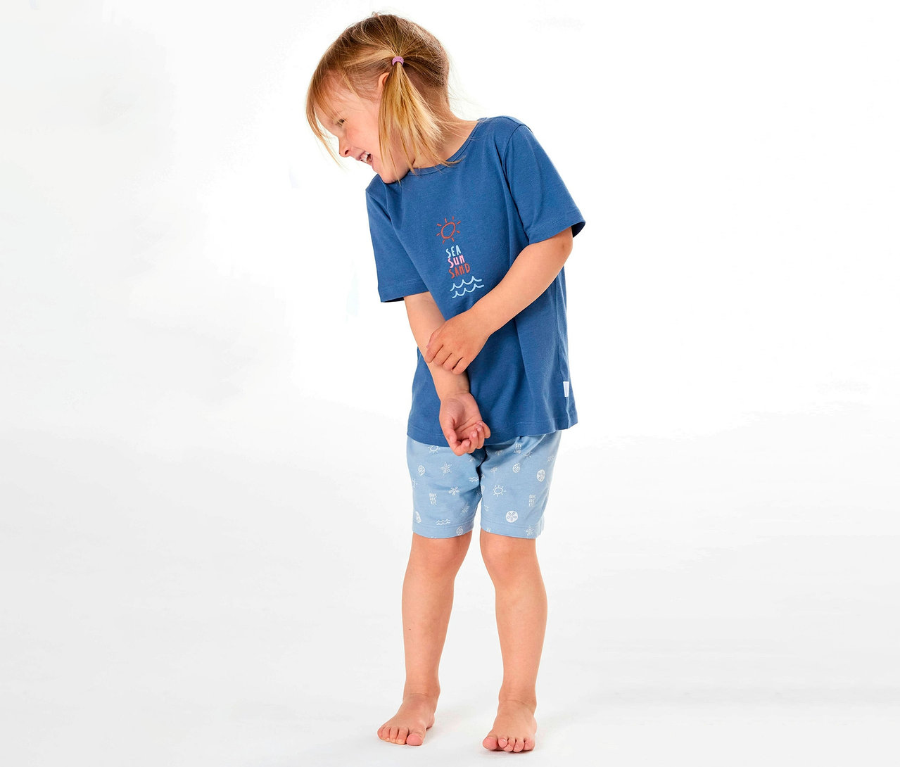 Une fille porte un pyjama court pour enfant SCHIESSER avec un t-shirt bleu et un short à motifs, souriant sur le côté.