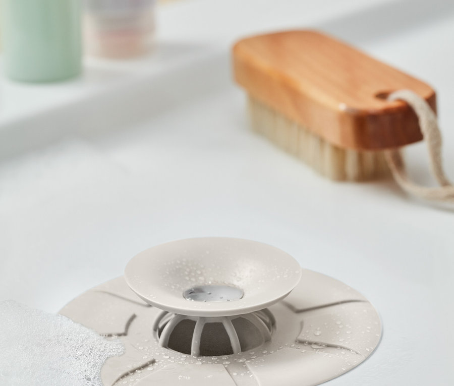 Un bouchon de lavabo beige avec un plateau repose sur un lavabo blanc, à côté se trouvent une brosse, une bouteille verte et un tube rose.