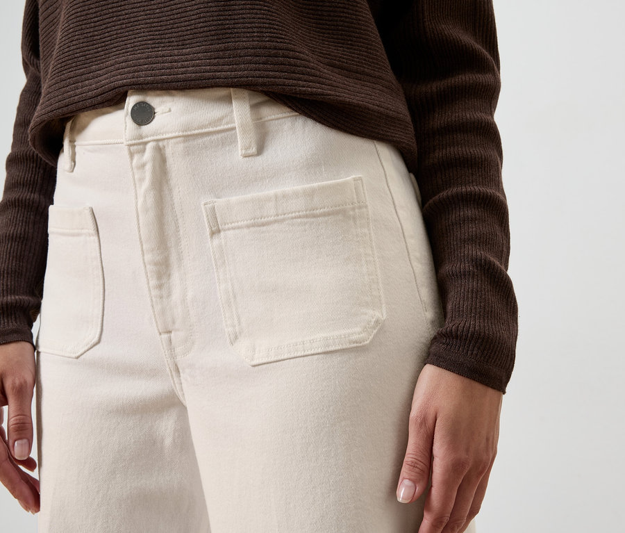 Gros plan d'une personne portant un pantalon crème avec des poches plaquées et un haut marron sur un fond neutre.