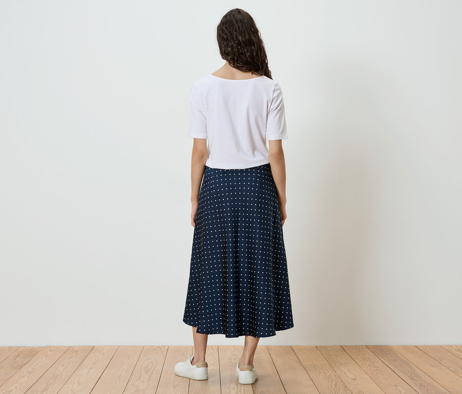 Vue de dos d'une femme portant un t-shirt blanc, une jupe bleue à pois blancs et des baskets blanches devant un mur blanc.