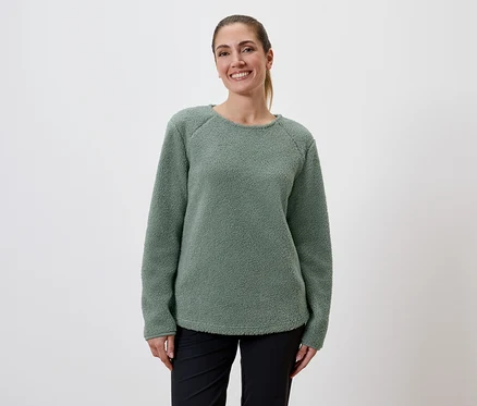 Une femme souriante avec une queue de cheval porte un pull en polaire vert et un pantalon noir sur un fond blanc.