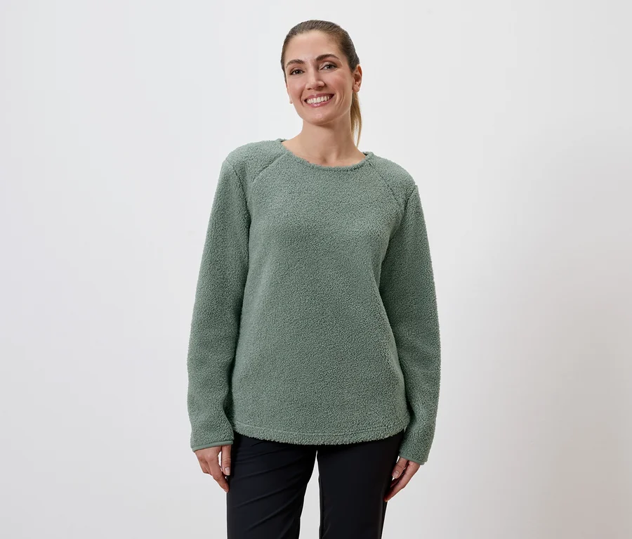 Une femme souriante avec une queue de cheval porte un pull en polaire vert et un pantalon noir sur un fond blanc.