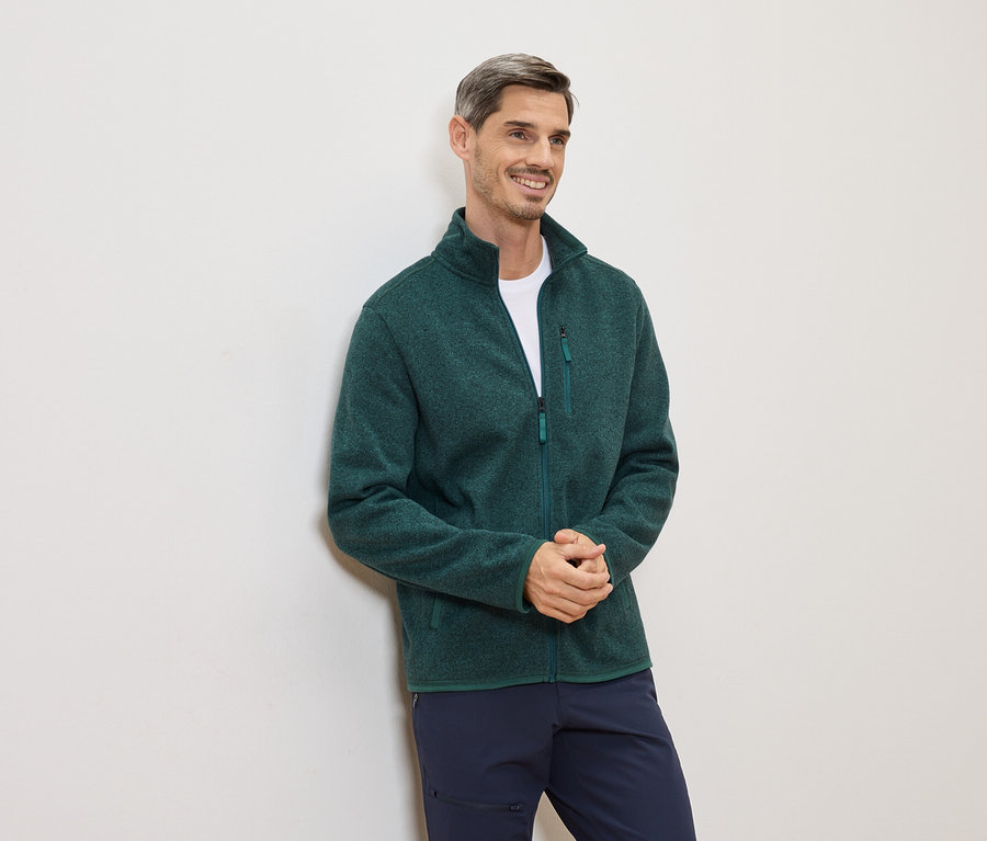 Un homme souriant portant une polaire verte et un pantalon bleu est appuyé contre un mur blanc.