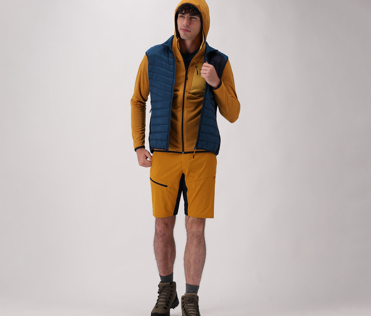 Un homme portant une veste à capuche CMP pour homme, un gilet hybride CMP pour homme, un short et des bottes sur fond clair.