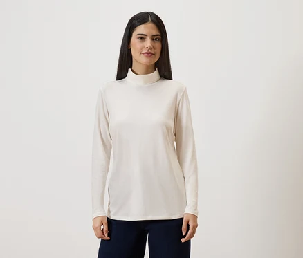 Une femme aux longs cheveux foncés porte un pull à col roulé crème à manches longues et un pantalon foncé sur un fond clair.