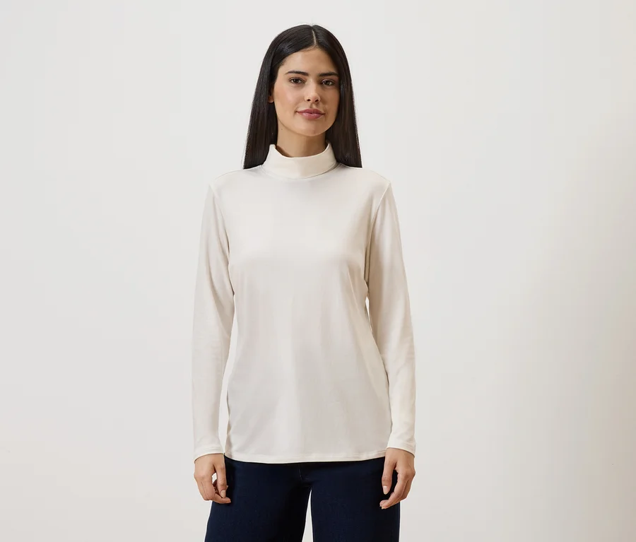 Une femme aux longs cheveux foncés porte un pull à col roulé crème à manches longues et un pantalon foncé sur un fond clair.