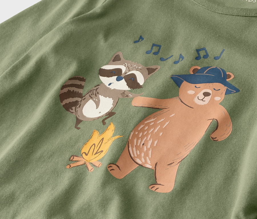Sur un tissu vert olive, un ours avec un chapeau et un raton laveur dansent à côté d'un feu de camp, accompagnés de notes de musique.