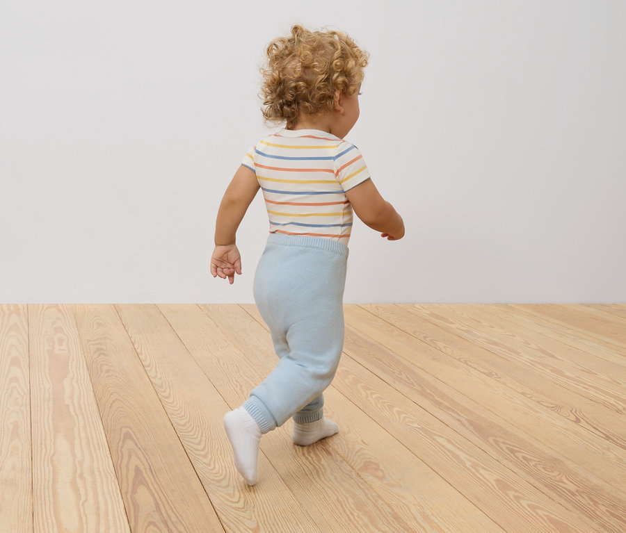 Un enfant aux cheveux bouclés, vu de dos, portant un body rayé et un pantalon bleu clair, debout sur un plancher en bois.