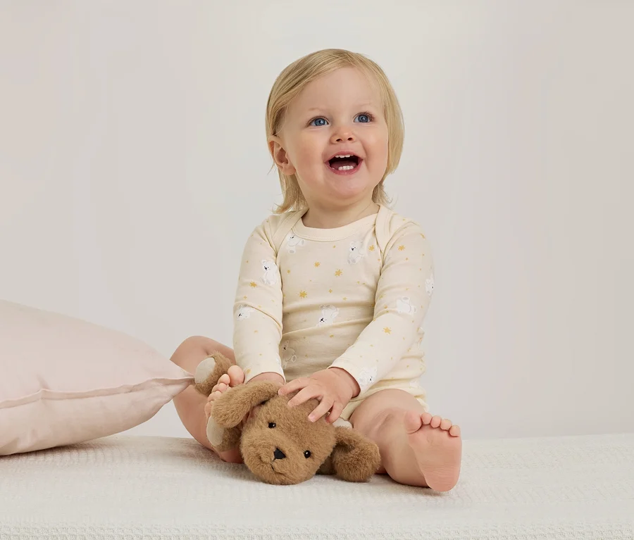 Un bébé souriant aux cheveux blonds est assis sur une couverture blanche, portant une grenouillère jaune clair à motifs d'ours et tenant une peluche marron. Un oreiller rose est à gauche.