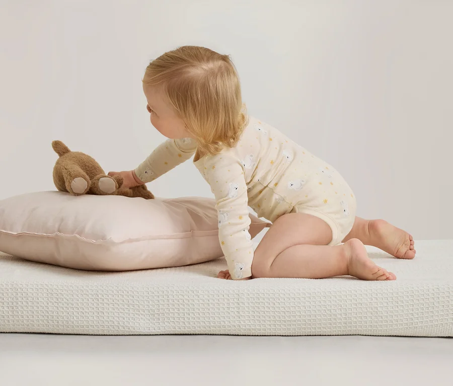 Un bébé en body à motifs est agenouillé sur une surface blanche texturée, tendant la main vers un ours en peluche marron posé sur un oreiller rose.