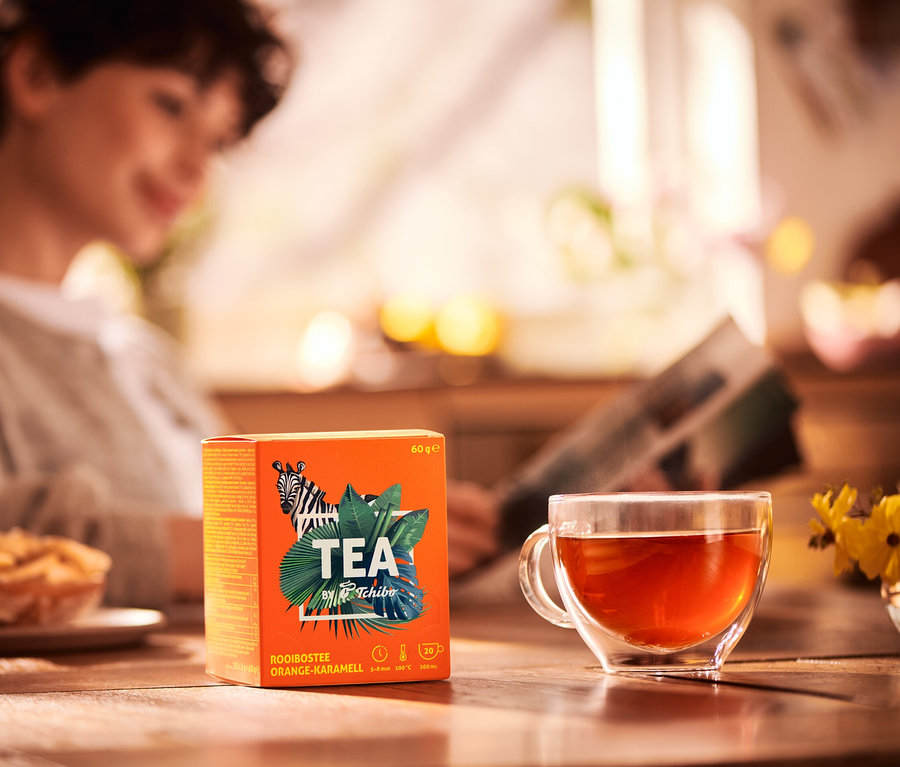 Femme lisant un journal à table avec une tasse de thé et un paquet de TEA by Tchibo Rooibos orange-caramel.