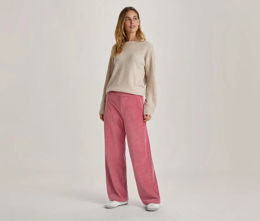 Une femme se tient debout, regardant vers la droite, vêtue d'un pull beige à col bateau, d'un pantalon large rose et de baskets blanches.