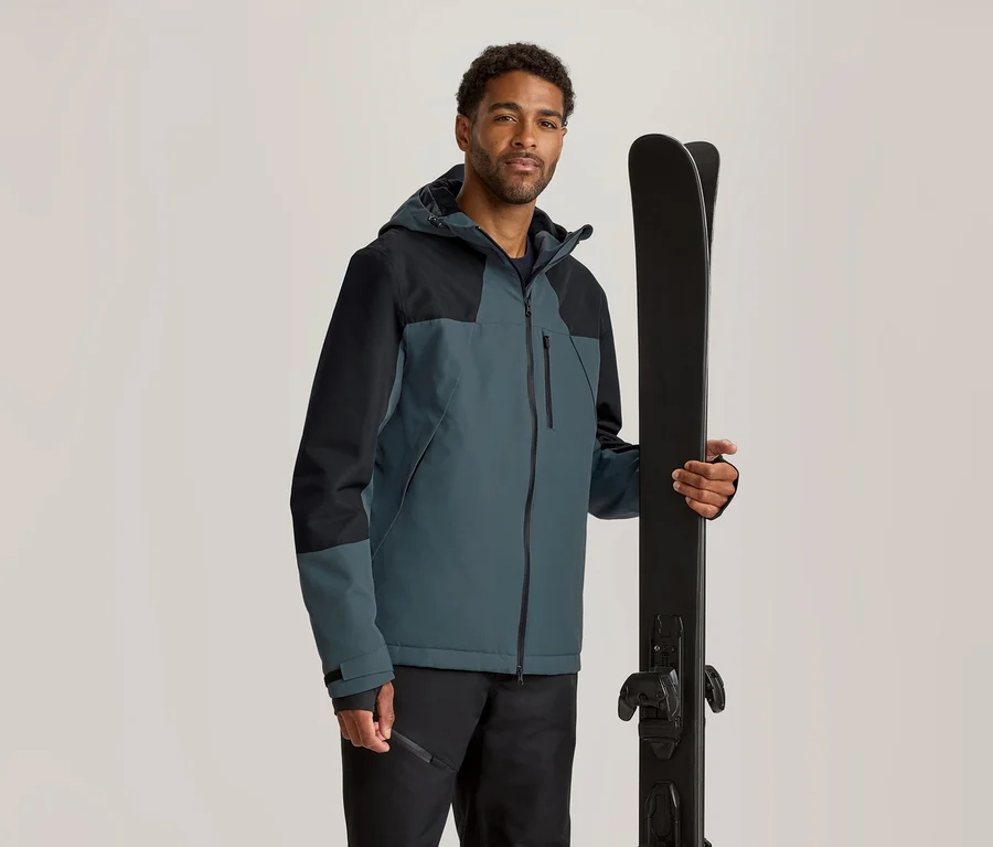 Homme portant une veste de ski bicolore bleu pétrole et noire, tenant des skis noirs verticalement.
