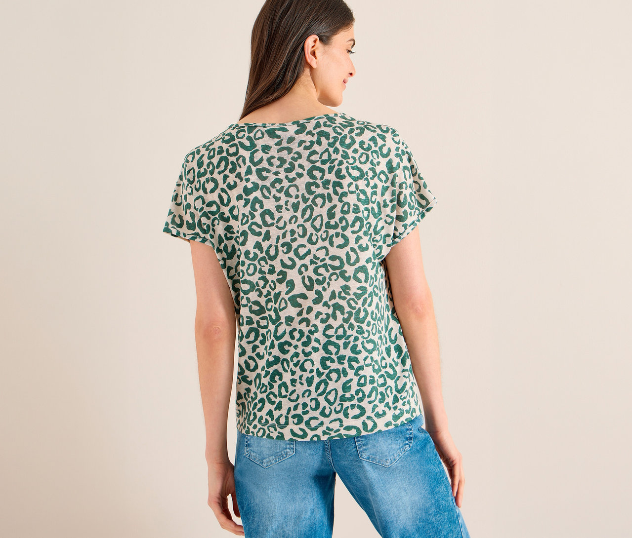 Une femme vue de dos, portant un T-shirt manches courtes CECIL beige à imprimé léopard vert et un jean bleu. Elle regarde vers sa droite.