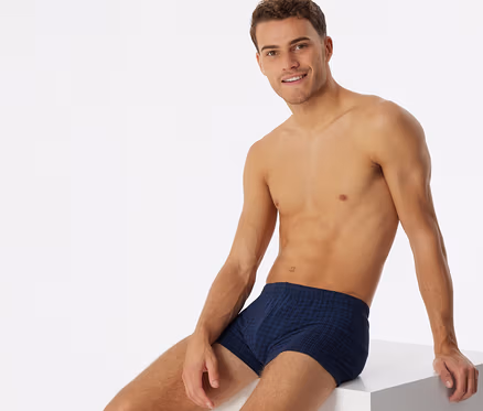 Homme torse nu, portant un short SCHIESSER à carreaux bleu foncé, assis et souriant sur un cube blanc.