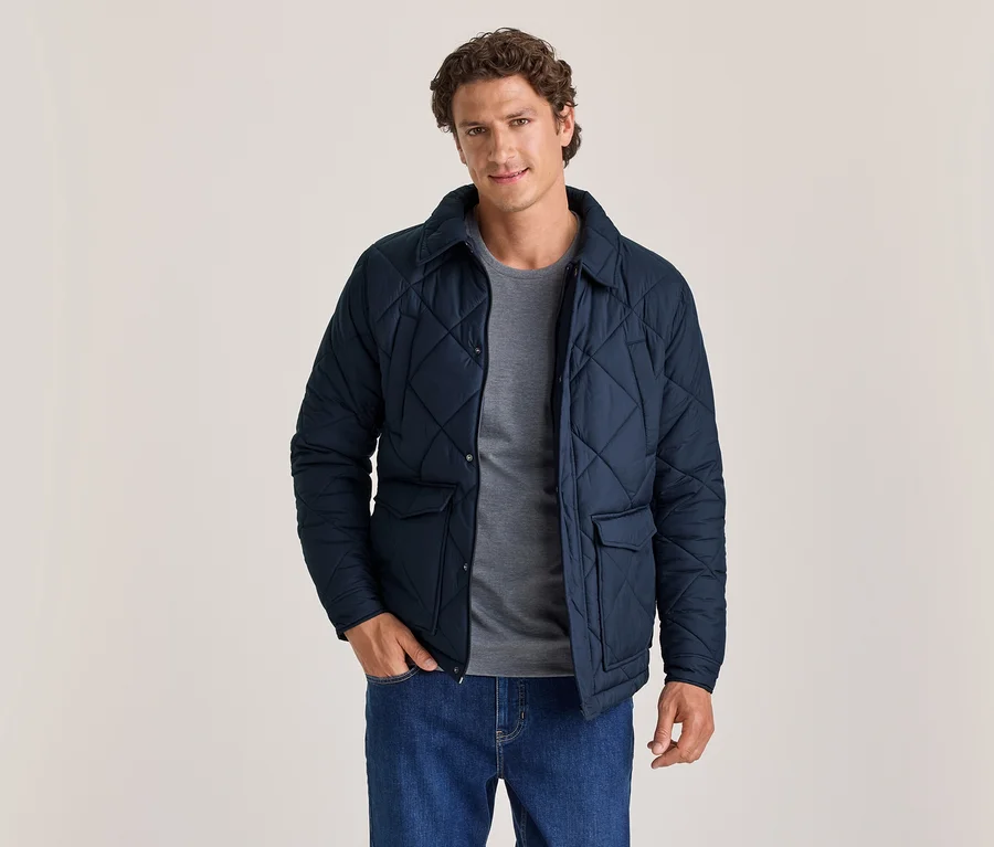 Homme portant une veste matelassée bleu marine sur un t-shirt gris et un jean bleu, debout, une main dans la poche, regardant l'appareil photo.